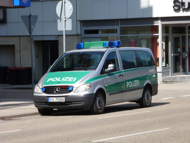 Ein Polizeiwagen steht auf der Seite der Straße vor einem Gebäude mit Glastüren, Schaufensterpuppen, Schildern und Mülltonnen im Hintergrund.