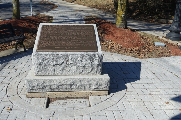 Ein steinernes Denkmal steht in einem Park, mit einer Bank links daneben, Bäumen, Pfählen und einer Straße im Hintergrund.
