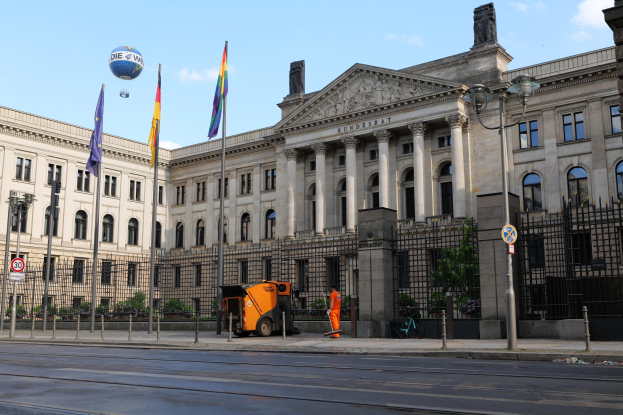 Großes modernes Gebäude mit vielen Fenstern, als Bundestag in Berlin bezeichnet, umgeben von Straßeninfrastruktur, Fußgängern, Flaggen und Bäumen unter einem bewölkten Himmel.