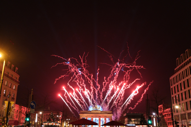 Eine belebte Stadtstraße an Silvester in Berlin, voller Menschen, Fahrzeuge und festlicher Dekoration, mit Feuerwerk, das den Nachthimmel erhellt.