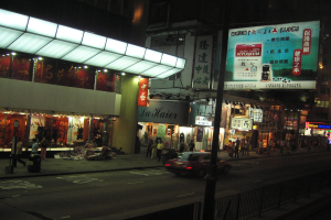 Eine nächtliche Stadtstraße mit einem vorbeifahrenden Auto, erleuchteten Gebäuden, Geschäftsfronten, Fußgängern auf dem Gehweg und sichtbaren Infrastruktur wie Strommasten und -kabel.