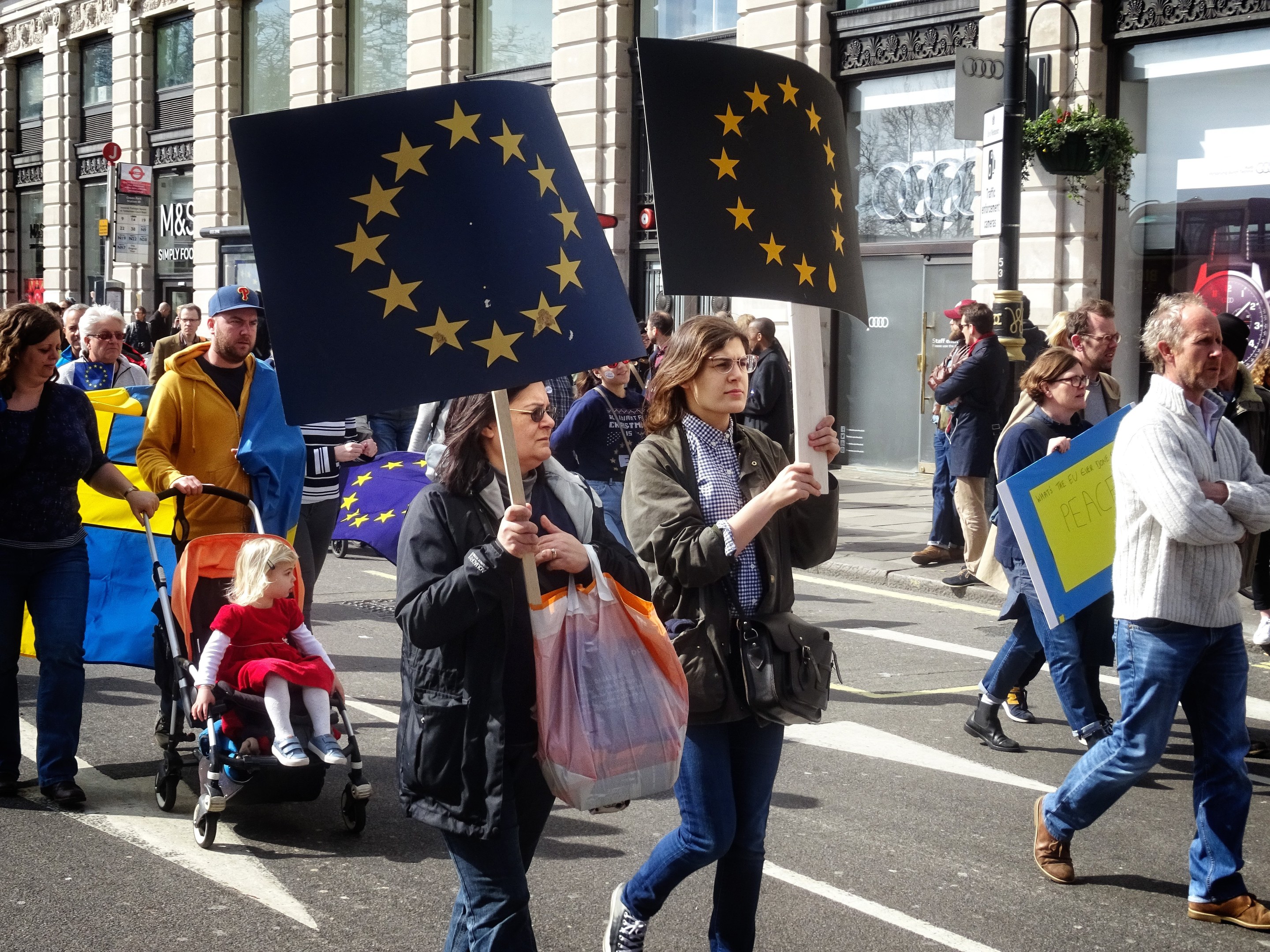 Menschen auf der Straße, einige mit Fahnen und Schildern, mit einem Baby im Kinderwagen und Gebäuden, Schildern und Topfpflanzen im Hintergrund.