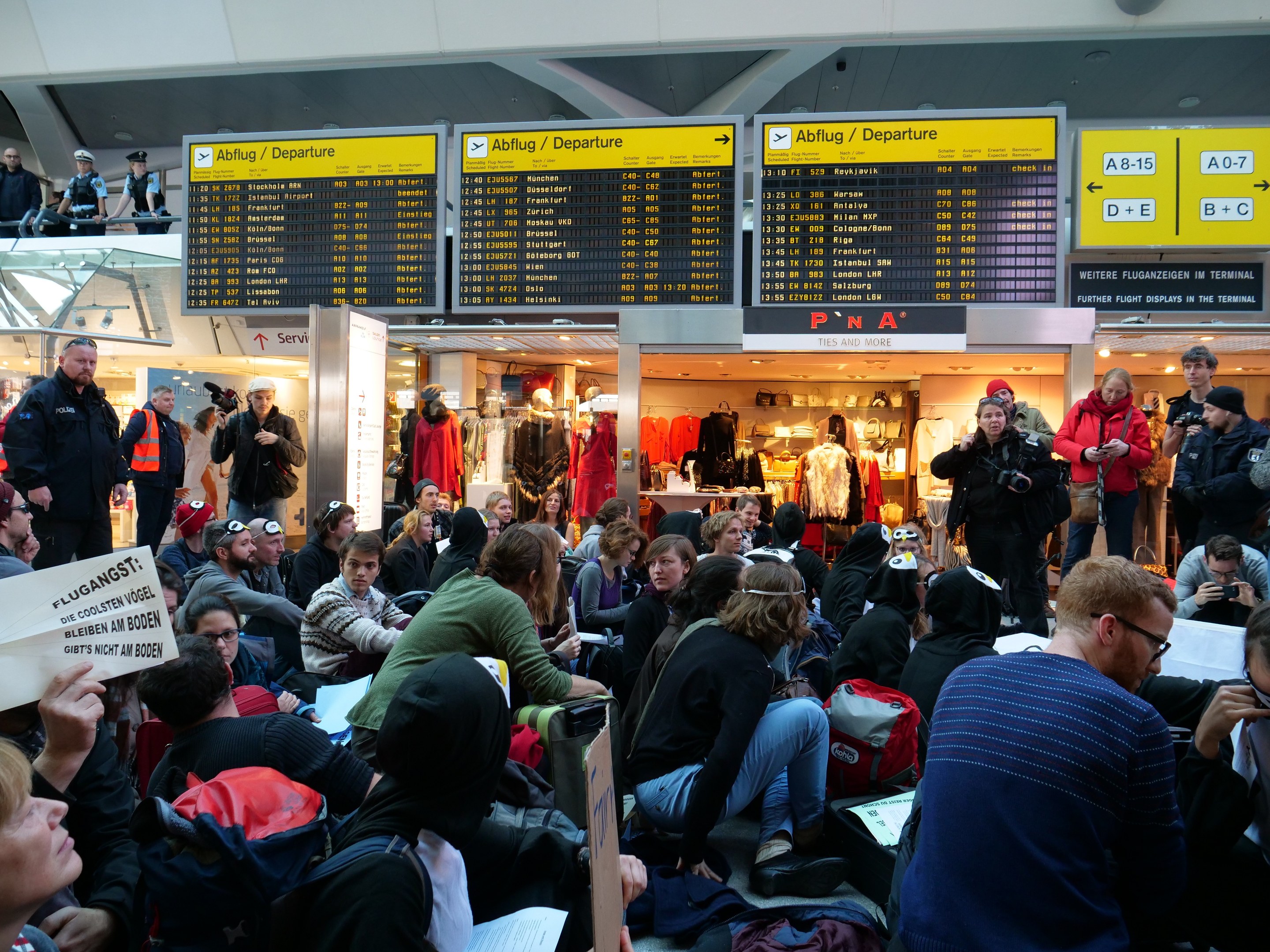 Eine große Gruppe von Menschen sitzt und steht in einem Flughafen während einer Protestaktion, wobei einige Taschen und Papiere halten, während Anzeigetafeln, Schaufensterpuppen in Kleidern und Deckenleuchten im Hintergrund zu sehen sind.