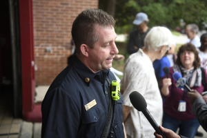 Ein Polizist in Uniform, der mit Reportern spricht, die Mikrofone halten, mit Bäumen und einem Gebäude im Hintergrund.
