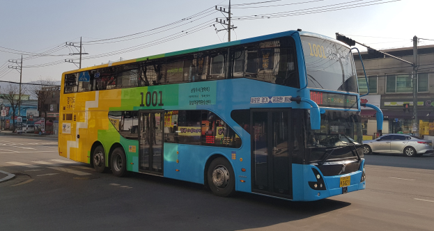 Blauer und gelber Doppeldeckerbus fährt auf einer Straße mit anderen Fahrzeugen, Strommasten, Gebäuden, Bäumen und einem klaren Himmel im Hintergrund.