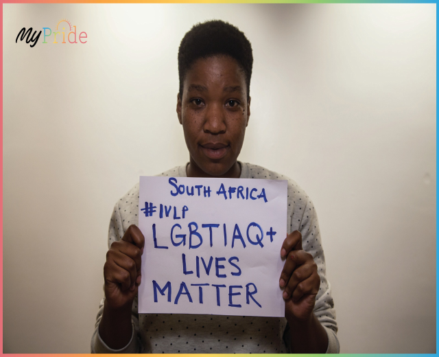 Eine Frau in einem weißen Hemd steht entschlossen vor einer Wand und hält ein Schild mit großer schwarzer Schrift, auf dem steht: "LGBTQ+ Lives Matter."