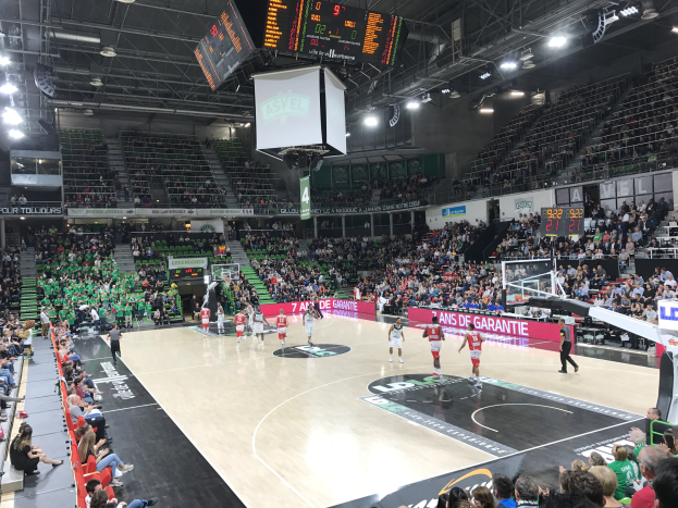 Basketballspiel in einer gro├čen Arena mit Publikum, Anzeigetafeln mit "Asvel vs Galatasaray" und überirdischen Lichtern und Bildschirm.