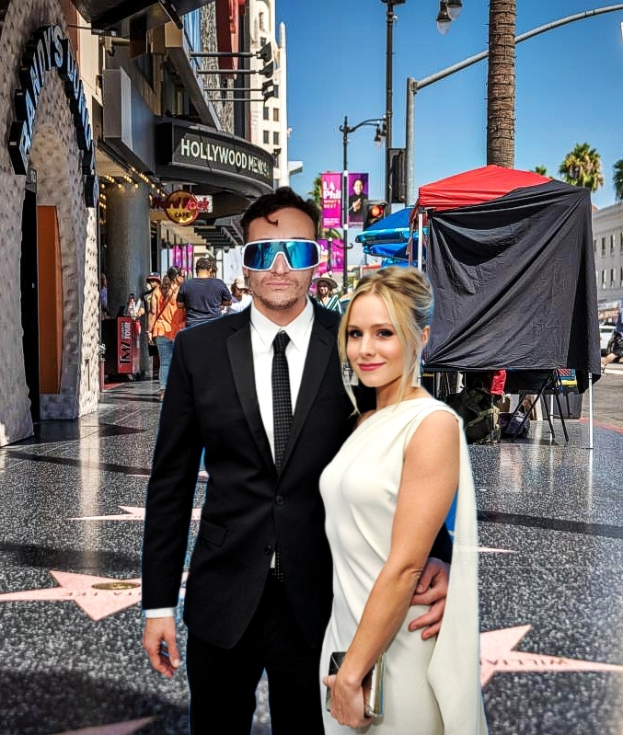 Ein Mann in einem schwarzen Blazer, weißem Hemd, schwarzer Krawatte und schwarzen Hosen steht neben einer Frau mit einer Geldbörse auf dem Hollywood Walk of Fame, mit Menschen, Gebäuden, Bäumen, Pfosten, Lichtern und Schildern im Hintergrund.