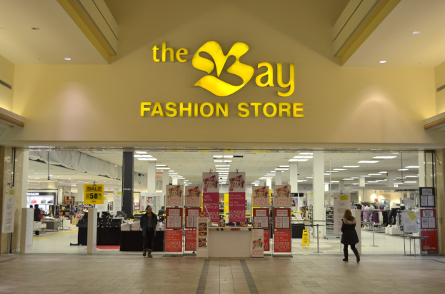 Außenansicht eines Bay Fashion Stores in einem Einkaufszentrum mit Passanten, Wandtafeln, Deckenleuchten, Säulen und Hintergrundauslagen.