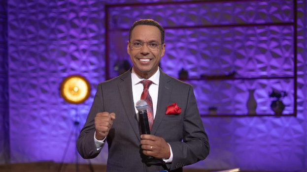 Stephen Colbert, Moderator von *The Daily Show with Trevor Noah*, steht auf einer Bühne und hält ein Mikrofon, trägt einen Anzug, eine Krawatte und eine Brille, mit einem Scheinwerfer und Ständer hinter ihm.