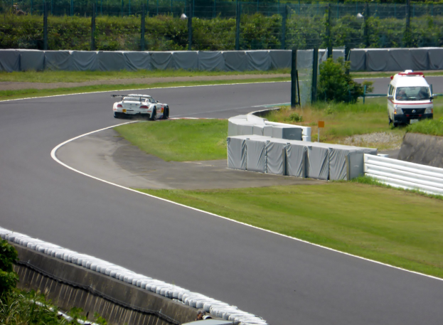 Ein Rennwagen, der auf einer Rennstrecke fährt, mit einem Polizeiwagen im Hintergrund, umgeben von Gras, Pflanzen, Bäumen und Absperrungen, mit einem Fahrzeug auf der rechten Seite und Text unten.