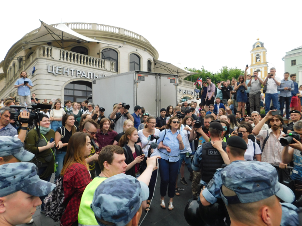 Eine große Gruppe von Menschen vor einem Gebäude stehend, einige mit Kameras und Mobiltelefonen, ein Fahrzeug in der Nähe geparkt, Bäume und ein klarer blauer Himmel im Hintergrund, scheinbar protestierend.