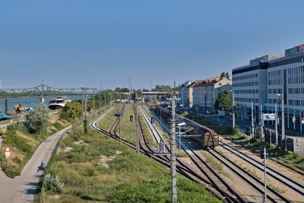 Ein Zug fährt auf Schienen neben einer Stadtlandschaft mit elektrischer Infrastruktur, Gebäuden, Grünflächen, Straßenschildern, Fahrzeugen, einer Brücke, Wasser und einem bewölkten Himmel.