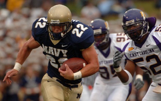 Ein Fußballspieler in einem marineblauen Trikot und Helm rennt mit dem Ball während eines Spiels, der Hintergrund ist unscharf, um die Geschwindigkeit anzuzeigen.