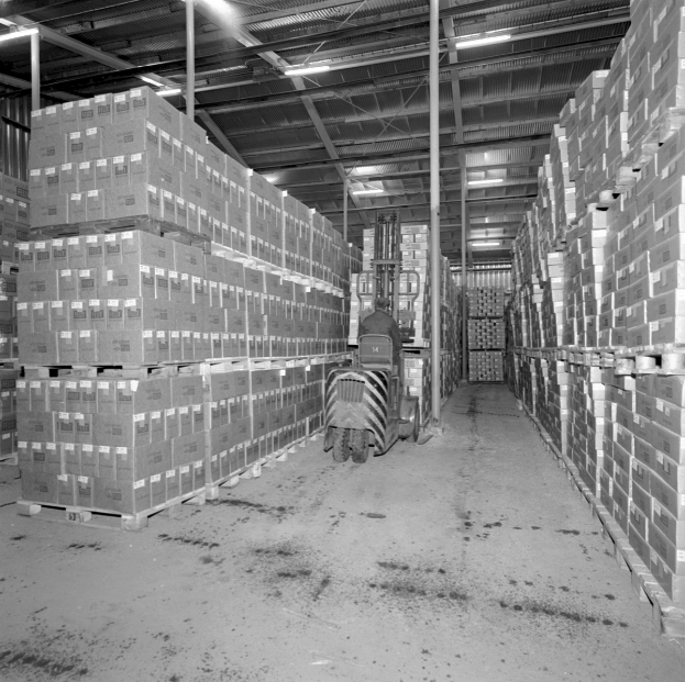 Ein Schwarz-Weiß-Foto eines großen Lagers mit vielen auf Regalen gestapelten Kisten und einem Fahrzeug in der Mitte, das von oben beleuchtet wird.