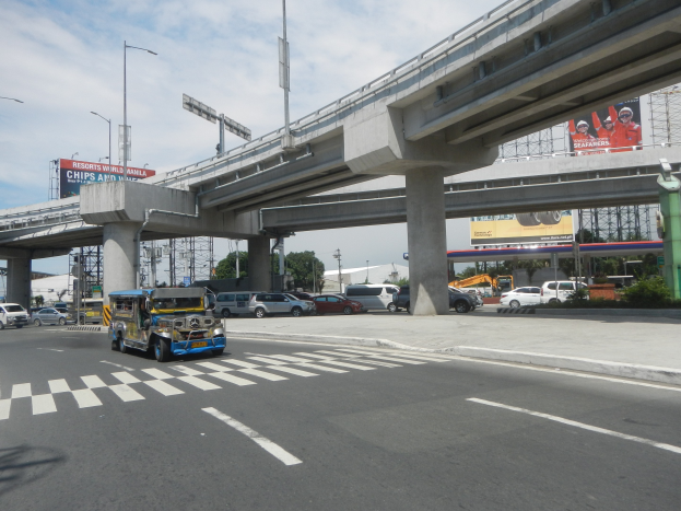 Vielbefahrene Hauptstraße mit Fahrzeugen, Zebrastreifen, Überführung mit Pfeilern, Laternen, Informationsschildern, Bäumen, Gebäuden und bewölktem Himmel.
