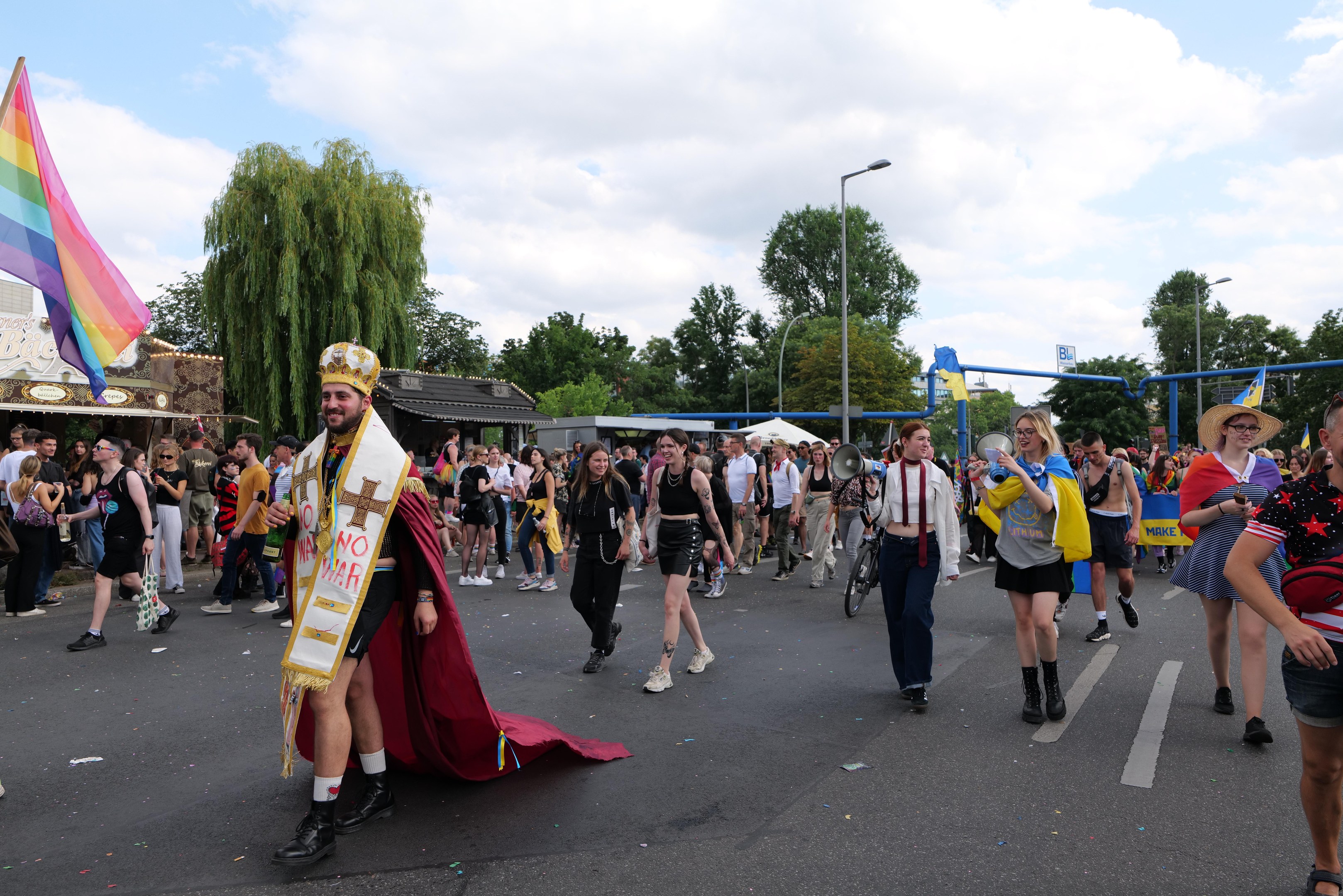 Eine Gruppe von Menschen marschiert auf der 2018 Gay Pride Parade mit einer Regenbogenflagge und Musikinstrumenten, einige tragen Mützen, vor einem Hintergrund aus Laternenmasten, Bäumen, Schuppen und einem bewölkten Himmel.