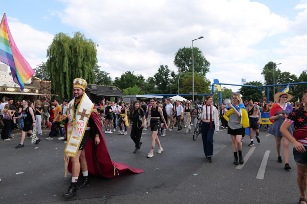 Eine Gruppe von Menschen marschiert auf der 2018 Gay Pride Parade mit einer Regenbogenflagge und Musikinstrumenten, einige tragen Mützen, vor einem Hintergrund aus Laternenmasten, Bäumen, Schuppen und einem bewölkten Himmel.