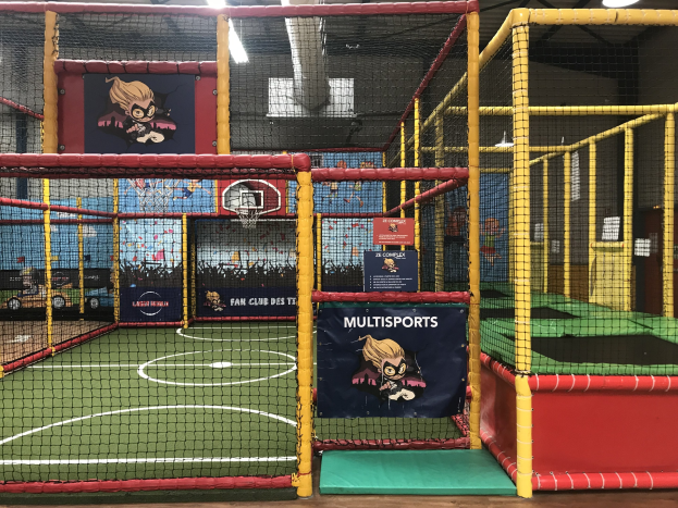 Großes Indoor-Trampolinpark mit einem Basketballfeld in der Mitte, umgeben von einem Netz, mit einem Basketballkorb, Wandbannern, Deckenbeleuchtung und sichtbarem Boden.