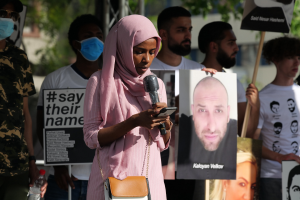 Eine Frau mit Kopftuch spricht in ein Mikrofon und hält ein Handy, während sie vor einer Gruppe von Menschen mit Schildern und einer Flasche steht, mit Bäumen und einem Gebäude im Hintergrund.