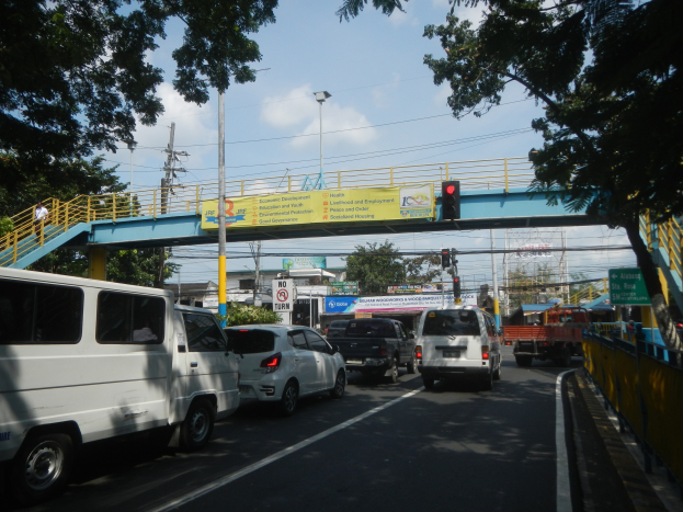 Eine belebte Straße mit Autos und Lastwagen, eine Brücke mit Geländern und Treppen, Laternen, Verkehrsampeln, Schilder mit Text, Bäume, Gebäude und ein bewölkter Himmel.