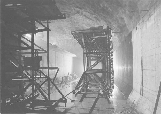 Schwarzes und weißes Foto eines Tunnels mit einer zentralen Metallstruktur, Metallstangen links, einer Wand rechts und Text unten.