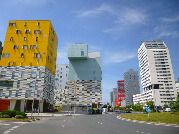 Eine Straße in einer Stadt mit hohen Gebäuden im Hintergrund, Straßenmäste, Straßenlaternen, Kraftfahrzeuge, Infotafeln, Büsche, Bäume und ein bewölkter Himmel, mit den einzigartig gestalteten Gebäuden der Universität Rotterdam.