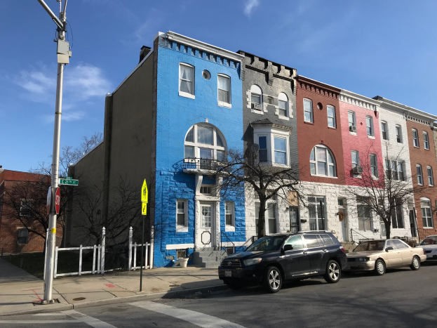 Eine Reihe bunter Häuser an einer Straßenecke mit parkenden Autos, Bäumen, Strommasten, einem Zaun und einem bewölkten Himmel in Baltimore, Maryland.