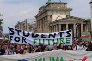 Eine Gruppe von Schülern marschiert in Berlin, hält ein buntes 'Students for Future'-Schild gegen eine Kulisse aus Gebäuden, Bäumen und Himmel.