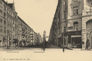 Schwarzes und weißes Foto von Charlottenburg, Deutschland, das eine Stadtstraße mit Gebäuden, Straßenlaternen, Lampen, Namensschildern, Kraftfahrzeugen, Fußgängern, Gittern und einem klaren Himmel zeigt.
