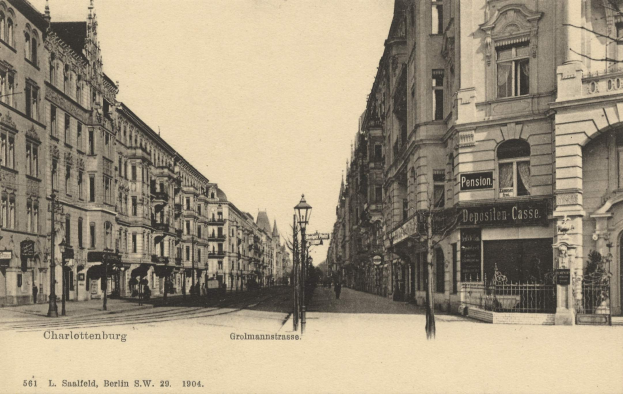 Schwarzes und weißes Foto von Charlottenburg, Deutschland, das eine Stadtstraße mit Gebäuden, Straßenlaternen, Lampen, Namensschildern, Kraftfahrzeugen, Fußgängern, Gittern und einem klaren Himmel zeigt.