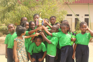 Gruppe von Kindern mit Pokal in einem Schulhof mit Bäumen, Pflanzen und einem Gebäude im Hintergrund