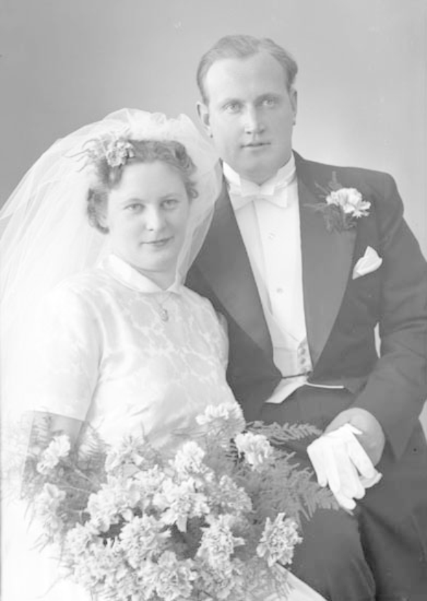 Ein Schwarz-Weiß-Foto eines Mannes und einer Frau an ihrem Hochzeitstag, die Frau hält einen Strauß Blumen in der Hand, beide lächeln und schauen sich an.