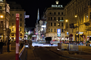Eine nächtliche Stadtstraße mit fahrenden Autos, Fußgängern auf den Gehwegen, parkenden Fahrrädern, Straßenlaternen, hohen Gebäuden, Absperrungen und einem sichtbaren Nachthimmel, mit dem Rolex-Logo auf einem Gebäude im Hintergrund.