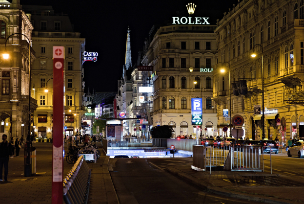 Eine nächtliche Stadtstraße mit fahrenden Autos, Fußgängern auf den Gehwegen, parkenden Fahrrädern, Straßenlaternen, hohen Gebäuden, Absperrungen und einem sichtbaren Nachthimmel, mit dem Rolex-Logo auf einem Gebäude im Hintergrund.