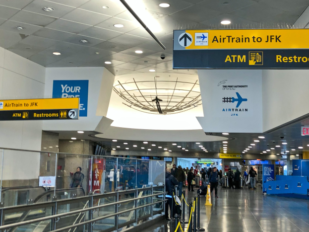 Eine Gruppe von Menschen geht durch eine Flughafenhalle mit einer Rolltreppe auf der linken Seite, Schilder an den Wänden, darunter eines mit der Aufschrift "Airtrain nach JFK", und Lampen an der Decke.