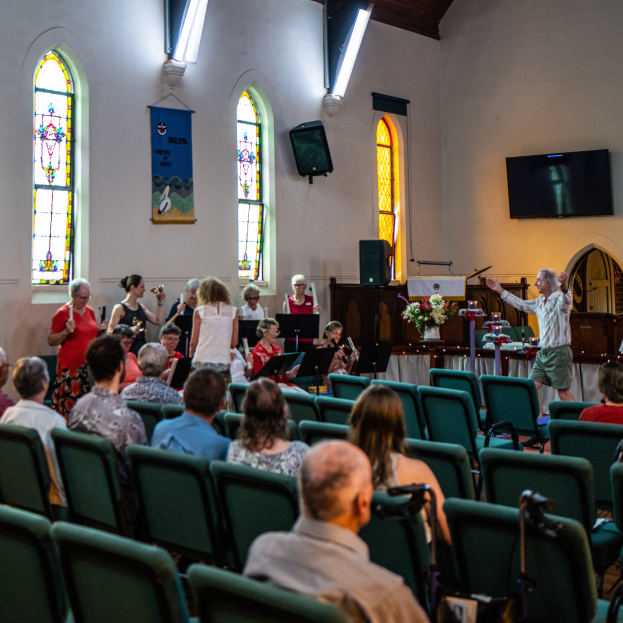 Eine Gruppe von Menschen sitzt auf Stühlen in einer Kirche, ein Mann steht mit einem Mikrofon vorne, umgeben von Musikinstrumenten, einem Tisch mit einer Blumenvase, einer Box, einem Banner mit Text an der Wand, Fenstern und Deckenleuchten.