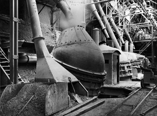 Ein Schwarz-Weiß-Foto einer großen Maschine in einer Fabrik, umgeben von verschiedenen Maschinen, Strukturen und Bahnschienen unten.