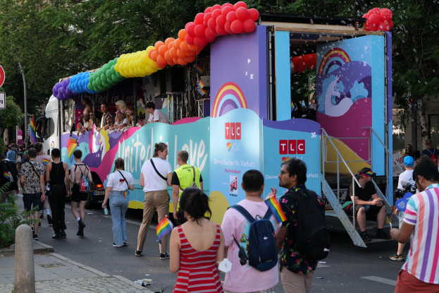 Eine Gruppe von Menschen, die auf einer Straße neben einem Lastwagen mit bunten Luftballons gehen, mit Schildern an Pfählen an der Straße und Bäumen und Gebäuden im Hintergrund, was auf eine Pride-Parade in Paris hindeutet.