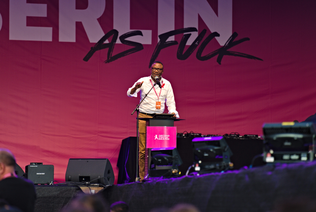 Mann in weißem Hemd und braunen Hosen hält Mikrofon auf Bühne mit pinkem Banner 'Berlin as Fuck' und Boxen