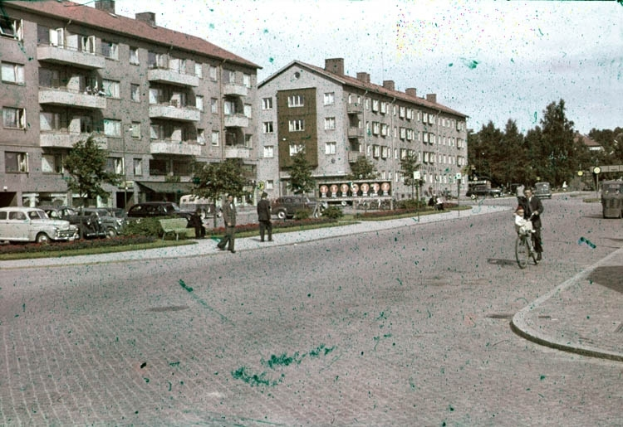 Ein altes Foto einer Stadtstraße mit geparkten Autos, einem Radfahrer im Vordergrund und Gebäuden, Bäumen und einem klaren blauen Himmel im Hintergrund.
