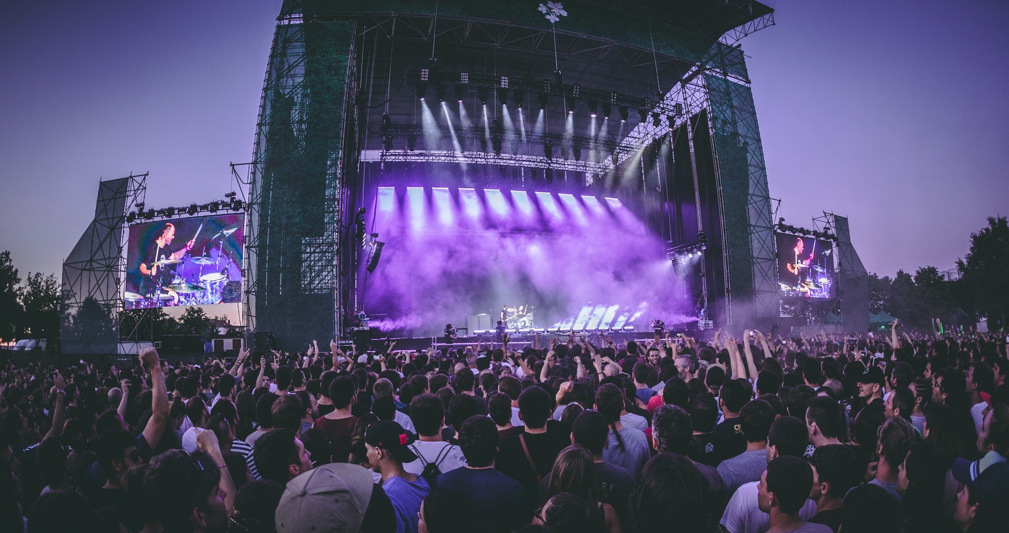 Eine große Menschenmenge bei einem Musikfestival schaut Künstlern auf einer hell erleuchteten Bühne mit Instrumenten und Bildschirmen zu, die vor einem Gebäude, Bäumen und einem blauen Himmel aufgebaut ist.
