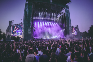 Eine große Menschenmenge bei einem Musikfestival schaut Künstlern auf einer hell erleuchteten Bühne mit Instrumenten und Bildschirmen zu, die vor einem Gebäude, Bäumen und einem blauen Himmel aufgebaut ist.
