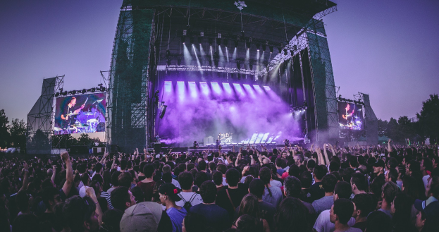 Eine große Menschenmenge bei einem Musikfestival schaut Künstlern auf einer hell erleuchteten Bühne mit Instrumenten und Bildschirmen zu, die vor einem Gebäude, Bäumen und einem blauen Himmel aufgebaut ist.