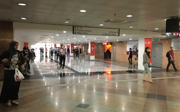 Menschen mit Taschen durch eine Flughafenhalle gehen, mit Hinweistafeln, Deckenlampen und einem Müllbehälter im Hintergrund; eine Frau trägt Schuhe.