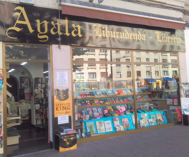 Verkaufsraum der 'Agala Liburudenda Libreria' mit Bücherregalen hinter Glas, Postern an der Wand und beleuchtet von Lampen, vor einer Stadtkulisse mit Gebäuden im Hintergrund.