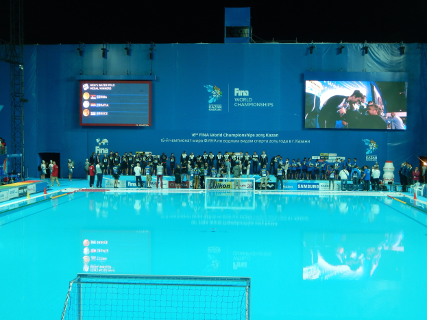 Gruppe von Menschen um einen Hallenbadpool mit Netz am Boden herumstehend, mit einer Wand mit "FINA World Championships 2015 Kazan"-Text und Logos, Trägern und Lichtern im Hintergrund.