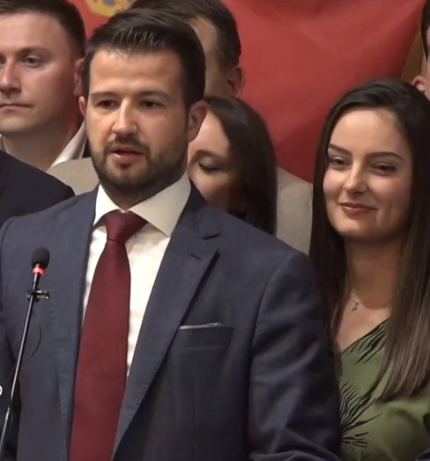 Ein Mann im Anzug spricht in ein Mikrofon an einem Rednerpult, mit einem Banner dahinter und einer Menge davor.