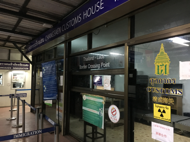 Der Eingang eines Zollamts in Bangkok, Thailand, mit Glaswänden und -türen, Informationsschildern mit Text und Bildern, Pfosten mit Bändern, einem Schild, einem Dach mit Deckenleuchten und einer Wand mit verschiedenen Gegenständen.