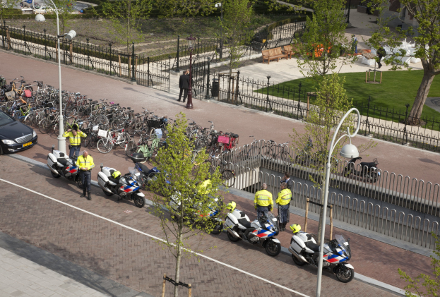 Eine Gruppe von Polizeibeamten steht neben geparkten Motorrädern am Straßenrand, mit einem geparkten Auto in der Nähe und einigen Menschen in der Nähe, während Bäume, Pfosten, Laternen, Geländer, Bänke, Gras und Gebäude im Hintergrund zu sehen sind.